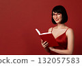 beautiful girl in a red dress and glasses reading a book against a red background 132057583