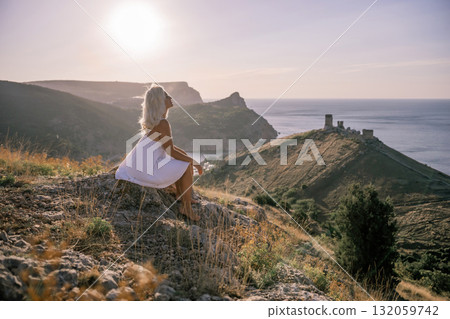 A woman is sitting on a hillside overlooking the ocean. She is wearing a white dress and has blonde hair. The scene is serene and peaceful, with the ocean in the background. A woman is sitting on a hillside overlooking the ocean. She is wearing a white dress and has blonde hair. The scene is serene and peaceful, with the ocean in the background. 132059742
