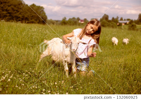 Girl cradling a baby goat in her arms, surrounded by a lush green field, basking in the warmth of a sunny day 132060925