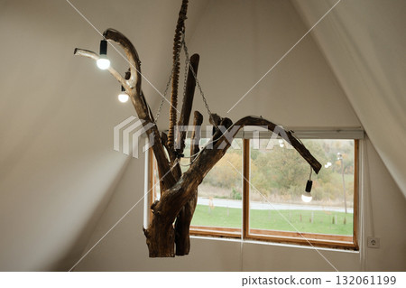 Artistic chandelier made from natural tree branches with hanging light bulbs, suspended in a bright attic interior. Perfect for rustic decor and design concepts 132061199