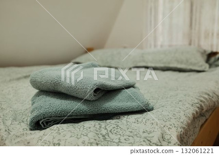 Folded soft towels placed on a neatly made bed with patterned linen and natural light through a lace curtain. Ideal for hospitality, home, or interior design use Folded soft towels placed on a neatly made bed with patterned linen and natural light through a lace curtain. Ideal for hospitality, home, or interior design use 132061221