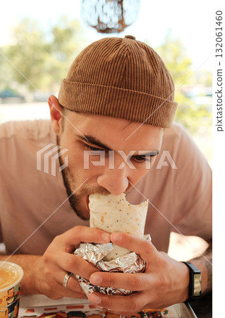 Man in a brown beanie eating a burrito wrapped in foil at a Mexican cafe table. Close front view portrait 132061460