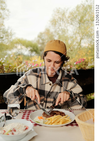 Woman in checkered jacket cutting grilled burger with fries and salad at outdoor restaurant during autumn day. Serbian traditional pljeskavica burger. Woman in checkered jacket cutting grilled burger with fries and salad at outdoor restaurant during autumn day. Serbian traditional pljeskavica burger. 132061512