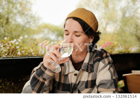 Woman smiling while holding and drinking a glass of water at an outdoor cafe table with flowers in background during autumn Woman smiling while holding and drinking a glass of water at an outdoor cafe table with flowers in background during autumn 132061514
