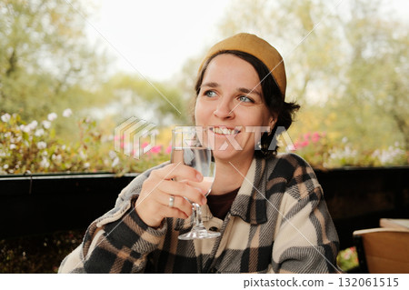 Woman smiling while holding and drinking a glass of water at an outdoor cafe table with flowers in background during autumn 132061515