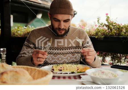 Man sitting at table with knife and fork looking at plate with traditional Balkan dish covered in cheese sauce, outdoor restaurant scene 132061526
