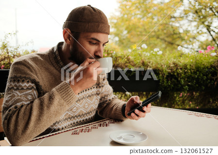 Young man in brown sweater holding coffee cup and using smartphone at outdoor cafe table, casual modern lifestyle scene 132061527