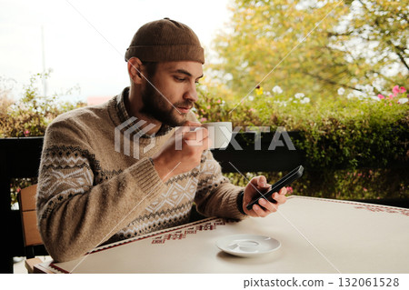 Young man in brown sweater holding coffee cup and using smartphone at outdoor cafe table, casual modern lifestyle scene Young man in brown sweater holding coffee cup and using smartphone at outdoor cafe table, casual modern lifestyle scene 132061528