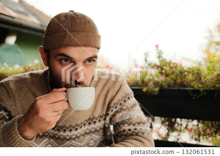 Man in warm sweater and hat drinking coffee at outdoor cafe table during autumn morning, calm and relaxed atmosphere 132061531
