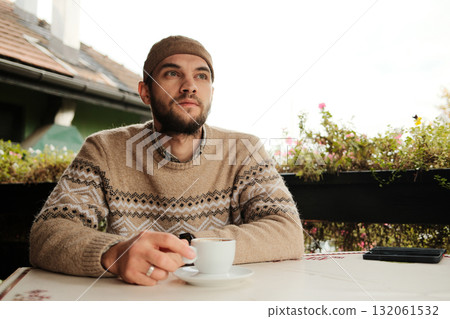 Man in warm sweater and hat drinking coffee at outdoor cafe table during autumn morning, calm and relaxed atmosphere 132061532