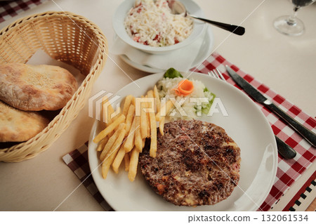 Plate with grilled Serbian burger - Pleskavica, french fries and onion served with bread and shopska salad in a white bowl on a red checkered tablecloth 132061534