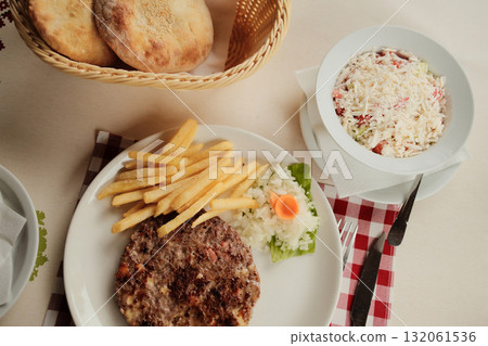 Plate with grilled Serbian burger - Pleskavica, french fries and onion served with bread and shopska salad in a white bowl on a red checkered tablecloth 132061536