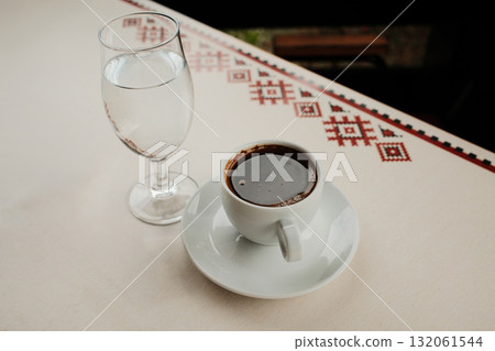 White porcelain cup with strong Turkish black coffee or americano and glass of water served on an outdoor cafe table in natural light 132061544