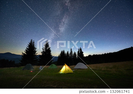 Several tents on grassy clearing under breathtaking, star-filled sky with Milky Way prominently visible. Pine trees and distant mountains frame scene, creating awe-inspiring camping experience. 132061741