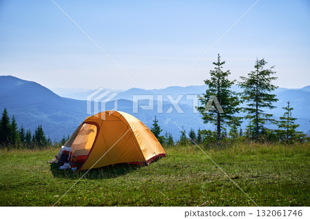 Bright yellow tent with camping gear on grassy hillside, overlooking breathtaking panorama of blue-tinged mountains. Young pine trees under clear blue sky, creating serene, picturesque camping spot. 132061746
