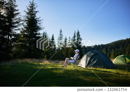 Woman camper relaxes next to tent with photo camera. Female tourist surrounded by tall evergreen trees and bathed in soft evening light, capturing peacefulness of moment. Woman camper relaxes next to tent with photo camera. Female tourist surrounded by tall evergreen trees and bathed in soft evening light, capturing peacefulness of moment. 132061754