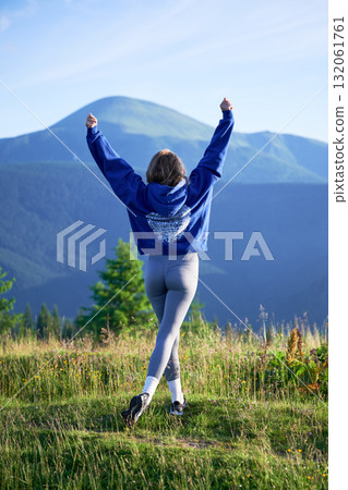 Woman tourist on grassy hill with beautiful mountain landscape in background. Young female wearing blue hoodie and grey leggings. 132061761
