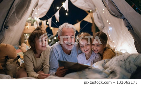 Grandfather reading a story to children in a cozy tent, surrounded by fairy lights, capturing the warmth of multigenerational family bonding 132062173