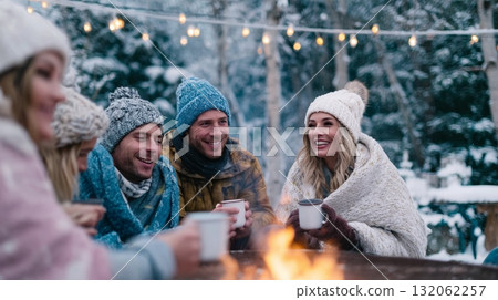 Group of friends gathered around a fire pit, enjoying warm drinks and cozy blankets during winter solstice celebrations in a snowy landscape 132062257