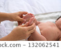 Mother hands gently cradle and massage tiny feet of newborn baby lying on white sheet. Infant baby wears white diaper on bed 132062541