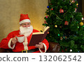Santa claus reading book and drinking hot beverage beside decorated christmas tree and twinkling lights in living room at home 132062542