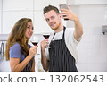 Happy couple taking selfie in modern kitchen. Man holding up smartphone to take picture while both holding cocktail glasses with red drink at home 132062543