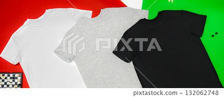 Close-up of the three t-shirts (black, white and grey) on color background redand green. Close-up of the three t-shirts (black, white and grey) on color background redand green. 132062748