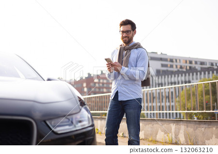 A man in glasses smiles while looking at his phone next to a black car outdoors. 132063210