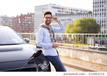 A man in glasses leans against a black car, holding a phone, with a city backdrop. A man in glasses leans against a black car, holding a phone, with a city backdrop. 132063214
