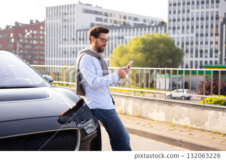 A man in glasses leans against a black car, engrossed in his phone, with a city backdrop. 132063226