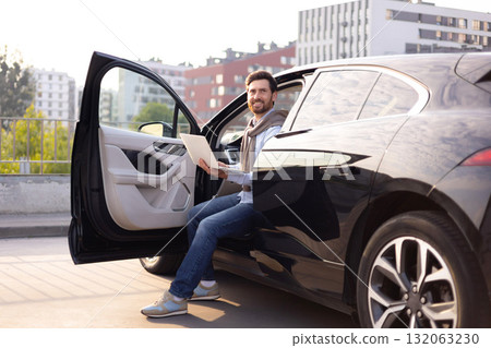 A man smiles while working on his laptop inside a black car with the door open, possibly on a rooftop parking area. A man smiles while working on his laptop inside a black car with the door open, possibly on a rooftop parking area. 132063230