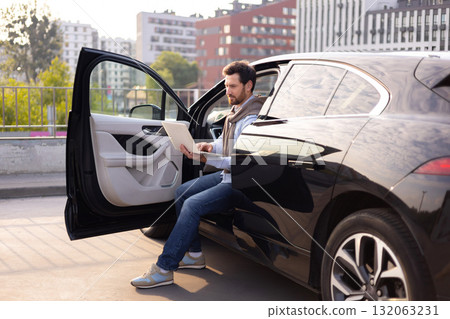 A man sits in a black car, working on a laptop with the door open, possibly in a parking lot. A man sits in a black car, working on a laptop with the door open, possibly in a parking lot. 132063231