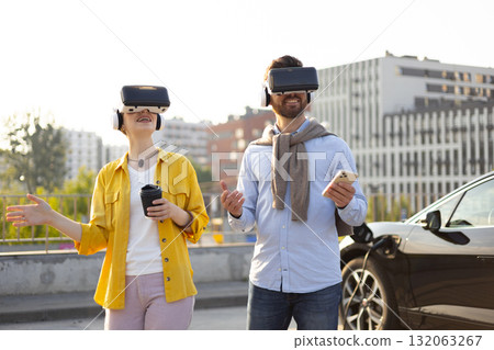A couple wearing VR headsets near an electric vehicle while it's charging, suggesting a futuristic experience. A couple wearing VR headsets near an electric vehicle while it's charging, suggesting a futuristic experience. 132063267