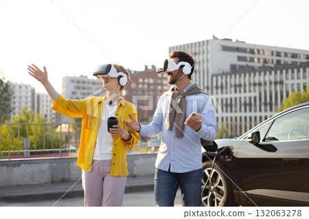 A couple wearing VR headsets near an electric vehicle charging, with one person gesturing towards the city. 132063278