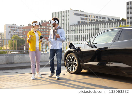 A couple wearing VR headsets chat near their electric vehicle while it charges. 132063282