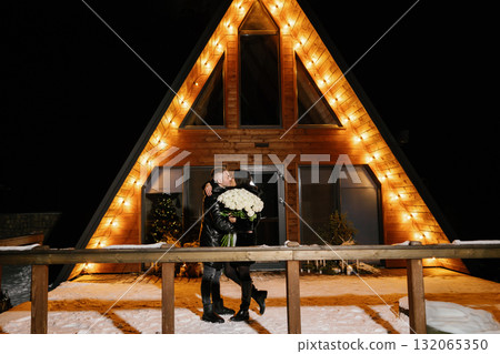Romantic couple hugging and holding white roses bouquet in front of their chalet during winter night 132065350