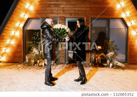 Romantic couple hugging and holding white roses bouquet in front of their chalet during winter night Romantic couple hugging and holding white roses bouquet in front of their chalet during winter night 132065352