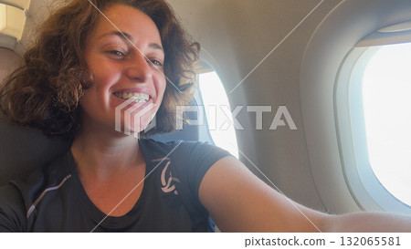 Happy passenger young woman is flying in plane. Smiling girl sitting in airplane near window going on trip vacation travel. Traveling female inside plane enjoying flight. Traveling girl 132065581