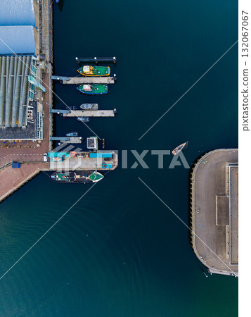Aerial view of harbor docks with moored boats, ridged-roof building, curved waterfront structure, and moving vessel in deep water, showcasing organized maritime infrastructure and dynamic contrast. Aerial view of harbor docks with moored boats, ridged-roof building, curved waterfront structure, and moving vessel in deep water, showcasing organized maritime infrastructure and dynamic contrast. 132067067
