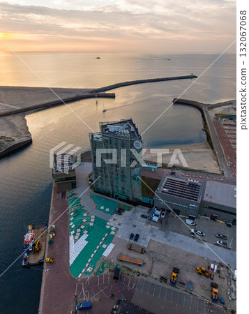 Aerial view of coastal harbor modern glass building near docks, construction equipment on site, breakwater protecting boats, sunset glow over sea blends urban design with maritime infrastructure. 132067068