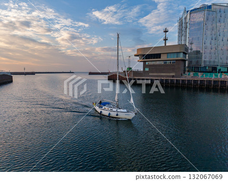Aerial view of harbor white sailboat with blue cover glides through calm water, modern glass building on dock reflects warm sunset light, blending serene motion with sleek architecture. 132067069