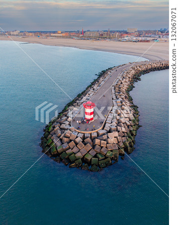 Aerial view of breakwater with red white lighthouse, people gathered near edge, concrete blocks protect harbor, coastal city and second tower in background under partly cloudy evening sky. Aerial view of breakwater with red white lighthouse, people gathered near edge, concrete blocks protect harbor, coastal city and second tower in background under partly cloudy evening sky. 132067071