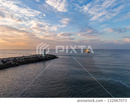 Aerial view of coastal scene rocky breakwater with greenwhite beacon, large yellow red ship sailing at sunrise or sunset, calm sea and warm sky tones create a tranquil maritime atmosphere. 132067073