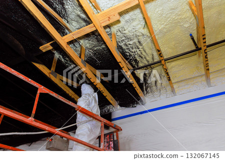 Contractor in protective gear applies refractory paint insulation using spray gun to wooden ceiling beams at construction site. 132067145