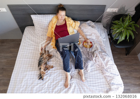 Woman Relaxing With Computer, Snacking, and Petting Dog in Bedroom 132068016