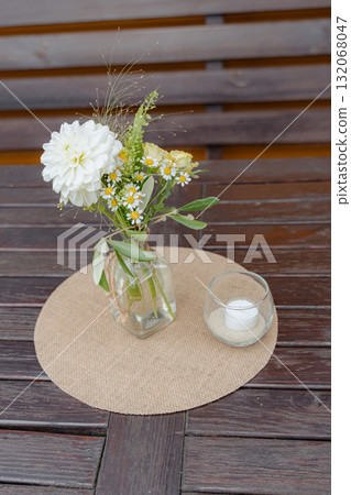 Charming floral arrangement with daisies and greenery in a glass vase, paired with a simple candle on a wooden table. Ideal for adding a touch of nature to interior decor Charming floral arrangement with daisies and greenery in a glass vase, paired with a simple candle on a wooden table. Ideal for adding a touch of nature to interior decor 132068047