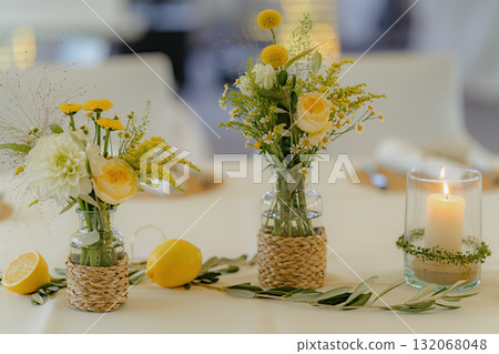 Beautiful table centerpiece featuring fresh yellow and white flowers in woven vases, lemons, and a lit candle in a glass holder. Perfect for weddings, events, or home decor elegance Beautiful table centerpiece featuring fresh yellow and white flowers in woven vases, lemons, and a lit candle in a glass holder. Perfect for weddings, events, or home decor elegance 132068048