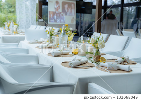 Wedding reception table elegantly set with floral arrangements and candles, creating a sophisticated atmosphere. Plates and cutlery neatly arranged enhance the visual appeal of the setup 132068049