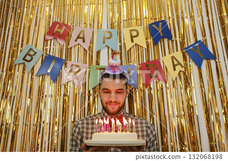 Man holding birthday cake with candles lit, golden backdrop and happy birthday banner behind Man holding birthday cake with candles lit, golden backdrop and happy birthday banner behind 132068198