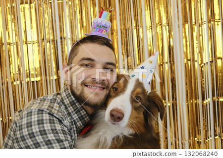 Man in party hat hugging Australian Shepherd dog at birthday celebration with golden background. Pet birthday party concept 132068240